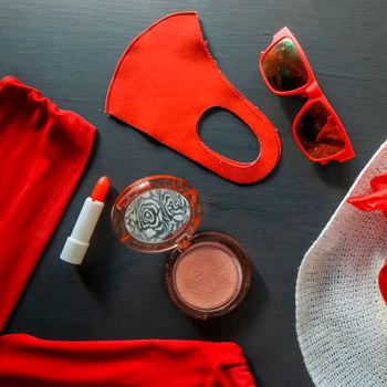 flat lay of a fashionable look from red and white accessories on a black background. A scarlet dress and a white hat with a ribbon, red lipstick, a fashionable medical mask, funny sunglasses.