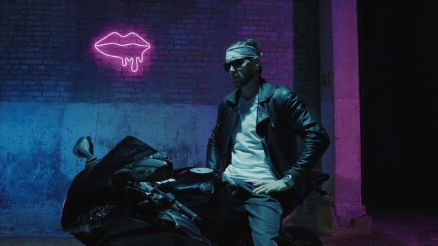 A man with a bandama sits on a sports motorcycle in sunglasses against a brick wall with a neon sign and smoke 4k