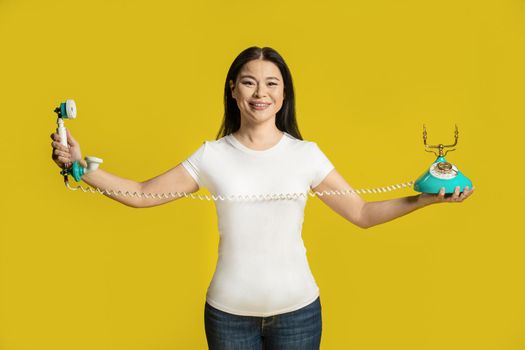 Stretching land line vintage phone cable funny asian woman hold telephone in hands wearing white t-shirt isolated on yellow background. Excited asian woman with retro phone. Communication concept.