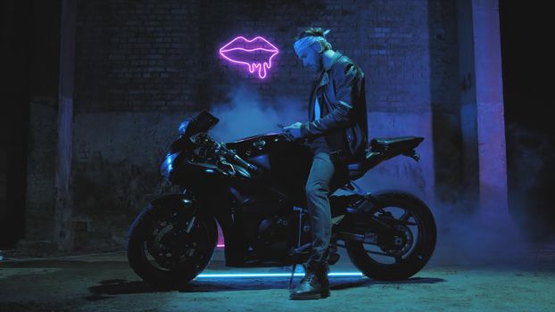 A man with a bandama sits on a sports motorcycle and texts on a mobile phone against a brick wall with a neon sign and smoke 4k