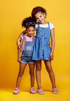 Studio portrait two mixed race girl sisters wearing funky sunglasses Isolated against a yellow background. Cute hispanic children posing inside. Happy and carefree kids imagining being fashion models.