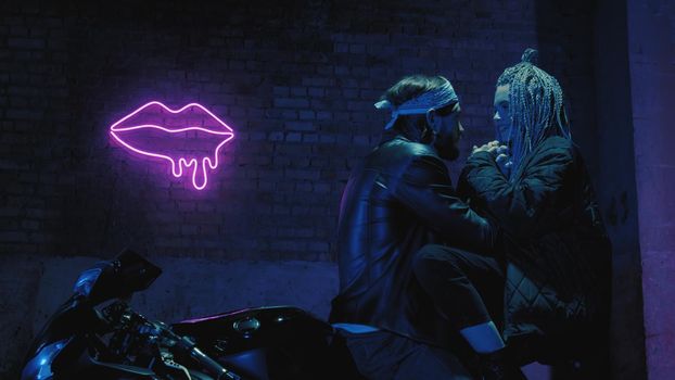 A girl in love and a guy are sitting on a super sport motorcycle flirting and hugging against the background of a neon sign 4k