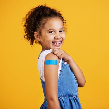 Studio portrait mixed race girl showing a plaster on her arm Isolated against a yellow background. Cute hispanic child lifting her sleeve to show injection site for covid or corona jab and vaccination.