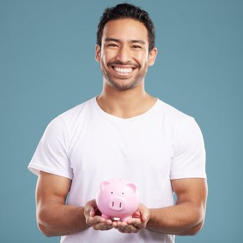 Handsome young mixed race man holding his pink piggybank while standing in studio isolated against a blue background. Hispanic male showing savings, finance, investment, wealth management and banking.
