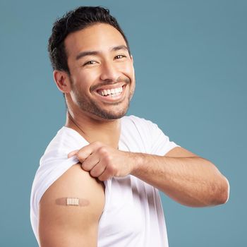 Handsome young mixed race man showing off his covid 19 jab while standing in studio isolated against a blue background. Hispanic male with a plaster on his corona virus vaccination injection site.