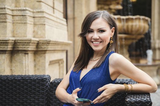 Elegant beautiful young woman with a smartphone in her hands looks at the camera while sitting in a street cafe.