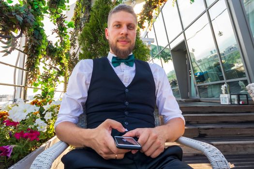 A young confident man in a white shirt, black vest with a smartphone in his hands looks at the camera while sitting in the penthouse.