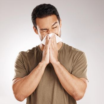 Handsome young mixed race man blowing his nose while standing in studio isolated against a grey background. Hispanic male suffering from cold, flu, sinus, hayfever or corona and using a facial tissue.