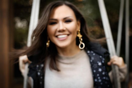 Blurred portrait of a young cheerful central asian woman swinging on a swing outdoor.