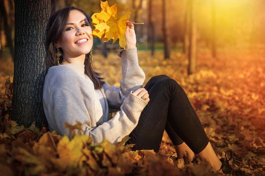 Young happy Central Asian woman sitting near tree and smiling at sunset in autumn park.