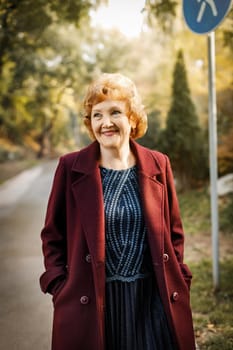 Russian mature woman with short hair smiling looks to the side outdoors in autumn in an unbuttoned burgundy coat. High quality photo