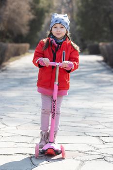 Happy preschool girl riding a scooter on a spring day in a public park.