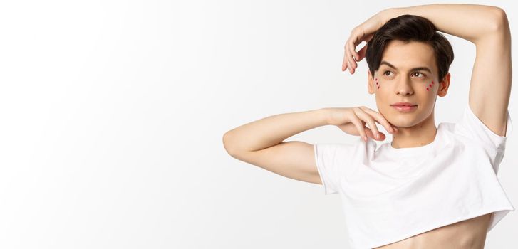 Close-up of beautiful androgynous man in crop top posing for camera, standing against white background.