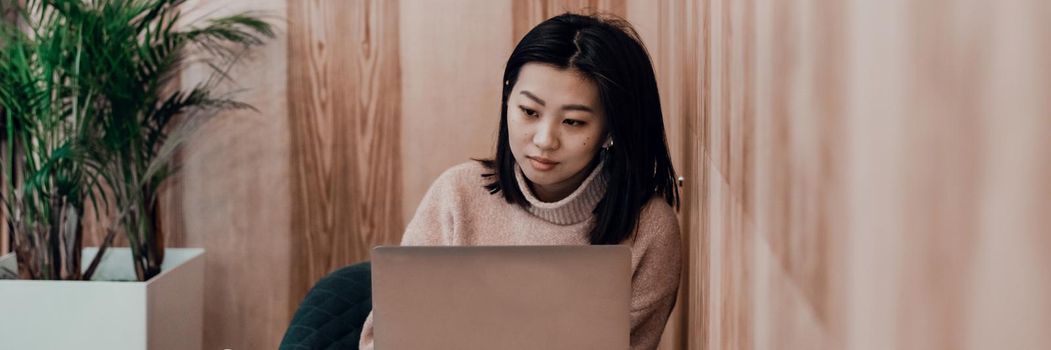 Cute adult girl plays in a laptop in a coffee shop. Asian woman working in a cafe at a laptop. Freelance concept. Web banner.