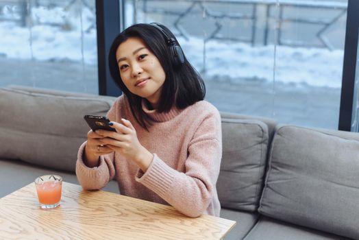 A cute Asian girl sits in a coffee shop, listens to music in large Bluetooth headphones and drinks freshly squeezed juice. Beautiful adult girl enjoys music in a public place and looks at the camera.