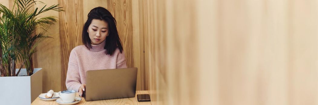 Beautiful asian girl sitting in a laptop in a cafe. Work in a coffee shop. Match and eclair next to a working laptop. Web banner.