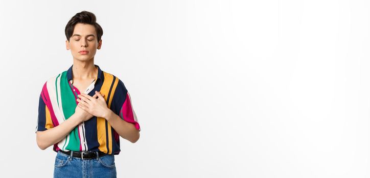 Image of nostalgic young gay man standing with closed eyes and hands on heart, remember something, standing over white background.