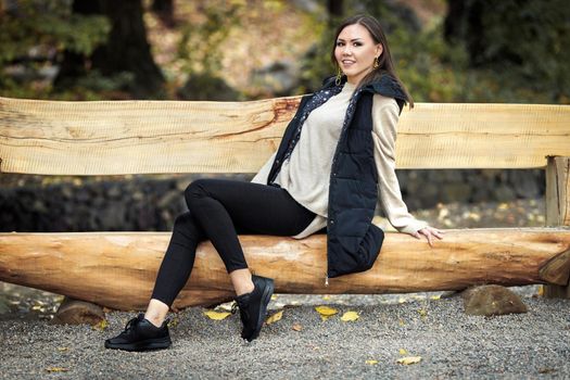 Autumn portrait of young asian woman with brown hair in basic colors clothing in park on wooden bench outdoors.