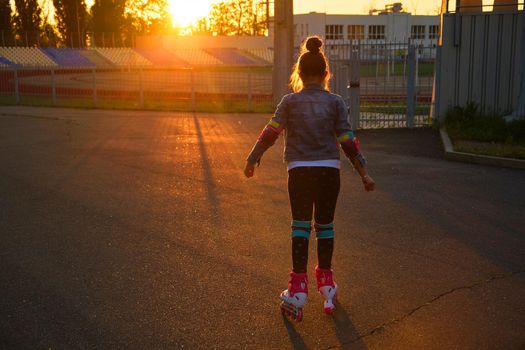 Little pretty happy funny girl on roller skates at stadium at sunset, learning to roller skate outdoors. Outdoor activity for children. Active sport for preschool kid. sun glare, selective focus
