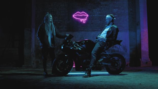 A guy on a super sport motorcycle stands talking to a girl against a pink neon sign 4k