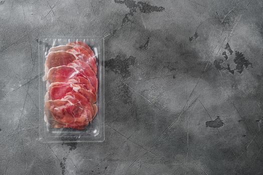 Prosciutto slices in transparent vacuum plastic pack set, on gray stone table background, top view flat lay, with copy space for text
