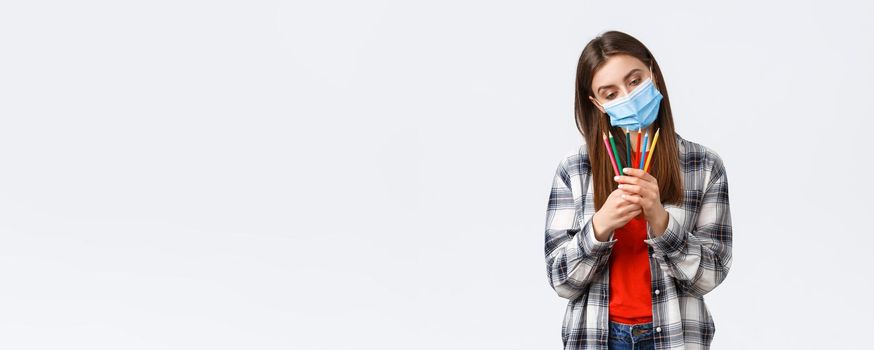 Social distancing, leisure and hobbies on covid-19 outbreak, coronavirus concept. Thoughtful cute, dreamy woman in medical mask decide which colored pencil choose, pondering what draw.