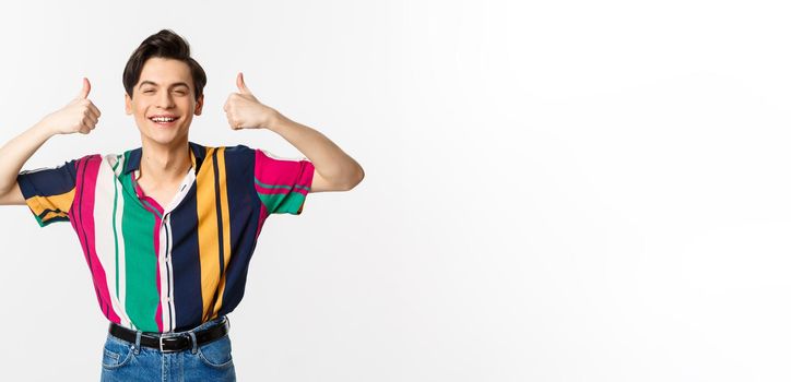 Young cheerful man smiling, showing thumbs up in approval, praise something good, standing over white background.