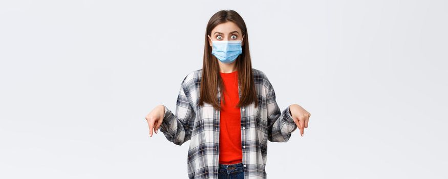 Coronavirus outbreak, leisure on quarantine, social distancing and emotions concept. Shocked and impressed young woman in medical mask and casual outfit, pointing fingers down, stare camera.