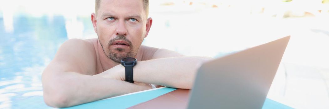 Annoyed man working on a laptop from the pool, close-up. Unexpected remote work while on vacation, distraction from rest