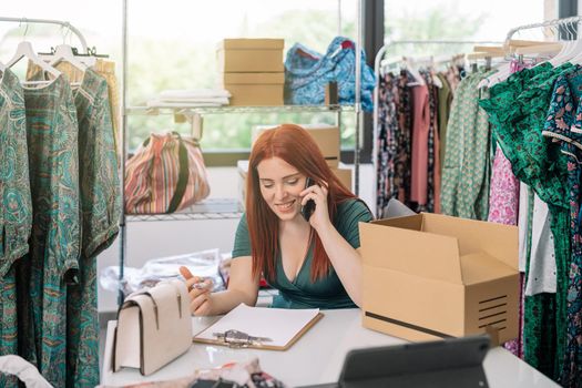 young woman businesswoman smiling, talking on her smartphone with her suppliers and customers in the warehouse of her clothing shop. work and business concept. natural light from window, background with clothes racks and clothes.