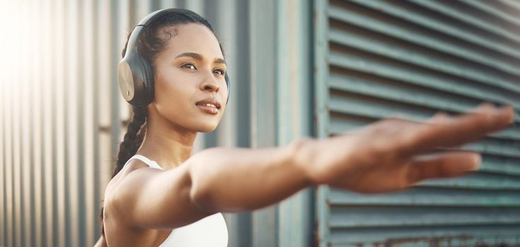 One fit young hispanic woman stretching arms in warrior pose for warmup to prevent injury while exercising in an urban setting outdoors. Focused and motivated female athlete listening to music with headphones while preparing body and mind for training workout or run.