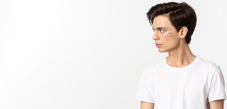 People, lgbtq community and lifestyle concept. Profile view of handsome gay man with glitter on face, looking left, standing over white background.