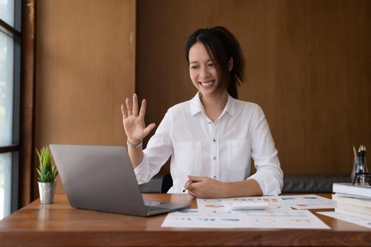 Young happy successful employee business asian woman waving hand sit work at workplace white desk talk by video call on laptop pc computer