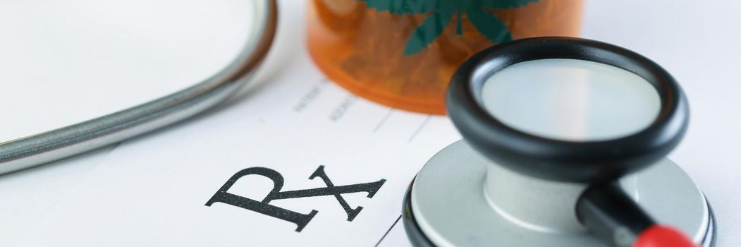 Close-up of plastic bottle with dried marijuana on blank medical prescription paper. Medication for patient, stethoscope tool. Alternative medicine concept