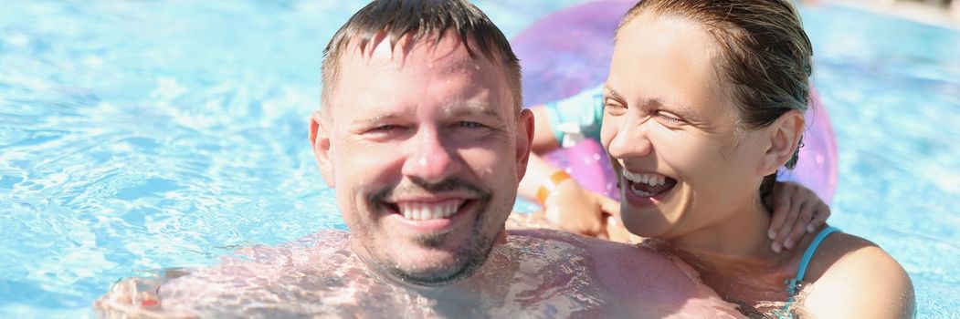 Portrait of cheerful couple posing for picture in swimming pool in luxury resort. Happy laughing woman and smiling husband hugging. Family, summer concept