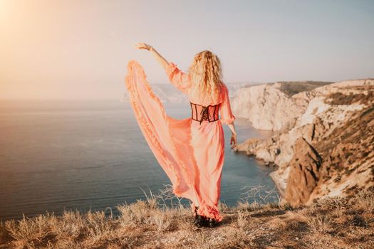 Side view a Young beautiful sensual woman in a red long dress posing on a volcanic rock high above the sea during sunset. Girl on the nature on blue sky background. Fashion photo