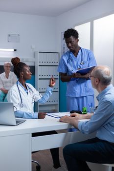 Hospital staff reviewing retired patient record file while talking about prescribed medicine. Clinic specialist discussing with nurse about consultation appointment dates.