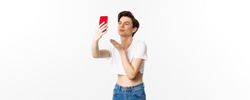 Beautiful gay man in crop top sending air kiss at phone camera, taking selfie or video chat on smartphone, standing over white background.