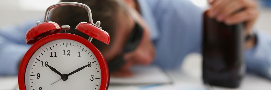 Close-up of red vintage clock show time, drunk man fell asleep on workplace in office. Worker hold alcoholic bottle, papers on table. Alcohol abuse concept