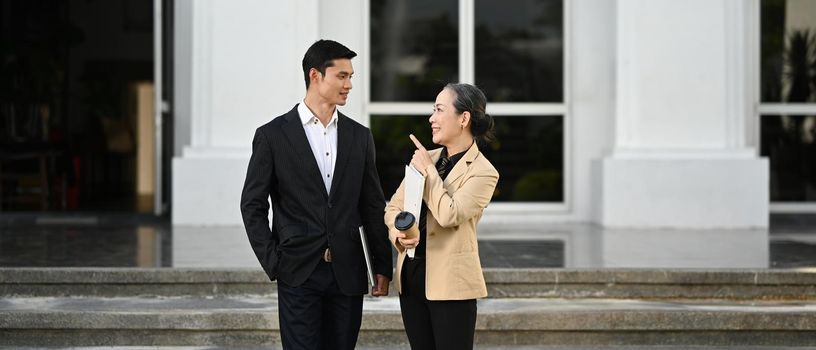 Two professional business people wearing formal suits talking and walking outside modern office building.
