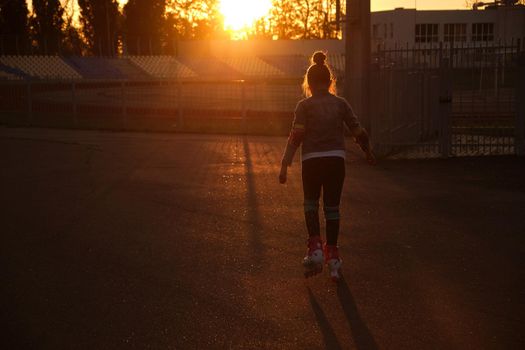 Little pretty happy funny girl on roller skates at stadium at sunset, learning to roller skate outdoors. Outdoor activity for children. Active sport for preschool kid. sun glare, selective focus