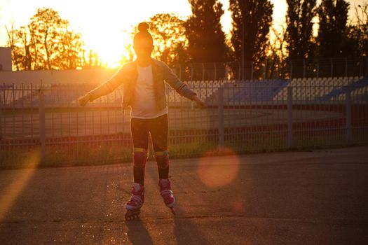 Little pretty happy funny girl on roller skates at stadium at sunset, learning to roller skate outdoors. Outdoor activity for children. Active sport for preschool kid. sun glare, selective focus