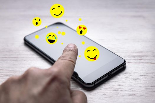 finger touching the screen of a phone that emoticons come out of on white wooden table, social media and communication concept, copy space for text, selective focus