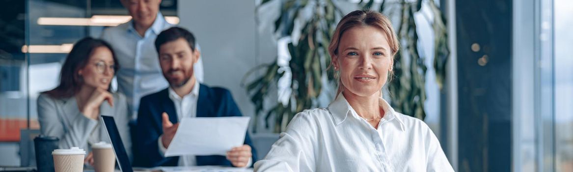 Smiling confident female boss sitting on meeting in office with her colleagues at background.