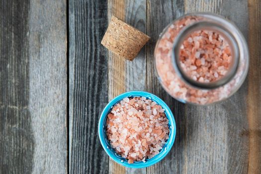 Pink Himalayan salt crystals in glass bottle and small bowl on wooden table flat lay.