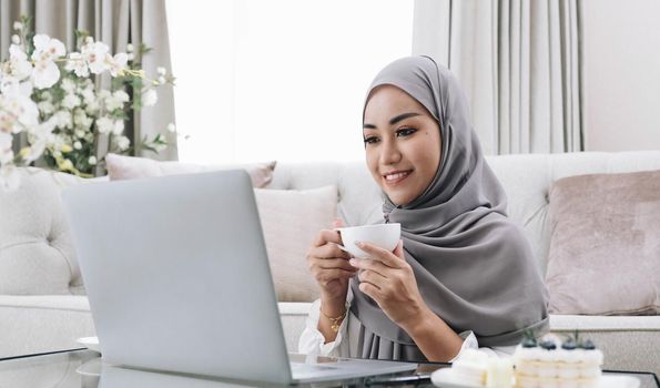 Domestic Lifestyle. Happy Muslim Woman Using Laptop At Home, Watching Movie And Drinking Coffee, Relaxing On Couch In Living Room, Side View.