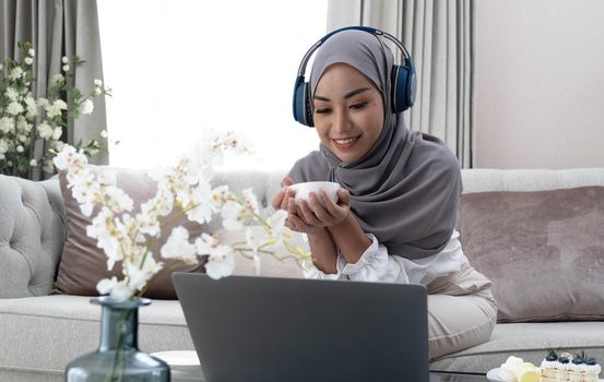 Domestic Lifestyle. Happy Muslim Woman Using Laptop At Home, Watching Movie And Drinking Coffee, Relaxing On Couch In Living Room, Side View.