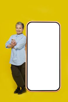 Leaned her back on huge, giant smartphone with white screen grey hair mature business woman with phone in hand looking at camera wearing casual isolated on white background. Free space mock up.