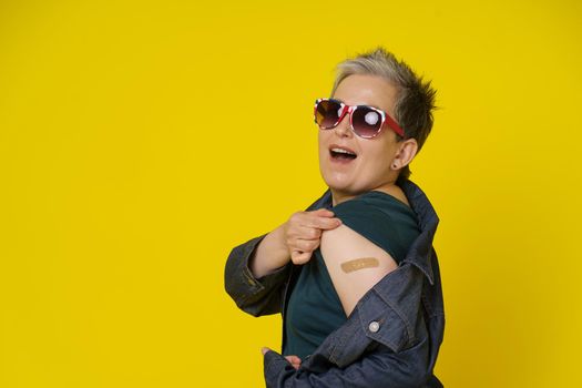 Senior woman with grey hair showing shoulder with band aid after having shot of vaccine wearing UK flag sunglasses isolated on yellow background. Mature woman 50s vaccination and healthcare concept.