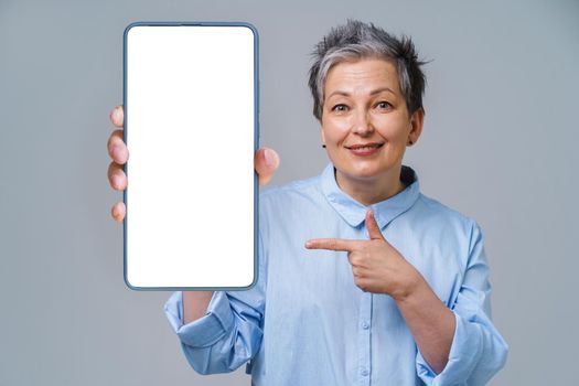 Mature silver, grey hair woman pointing at smartphone white screen holding it in her hand white screen mobile app advertising. Pretty woman in blue shirt isolated on white background.